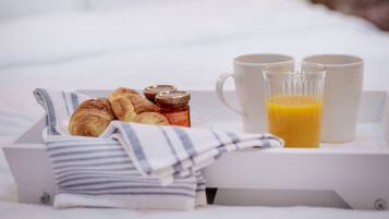 A neatly arranged breakfast tray with croissants, jam, and orange juice on a bed with white linens and a brick wall in the background.