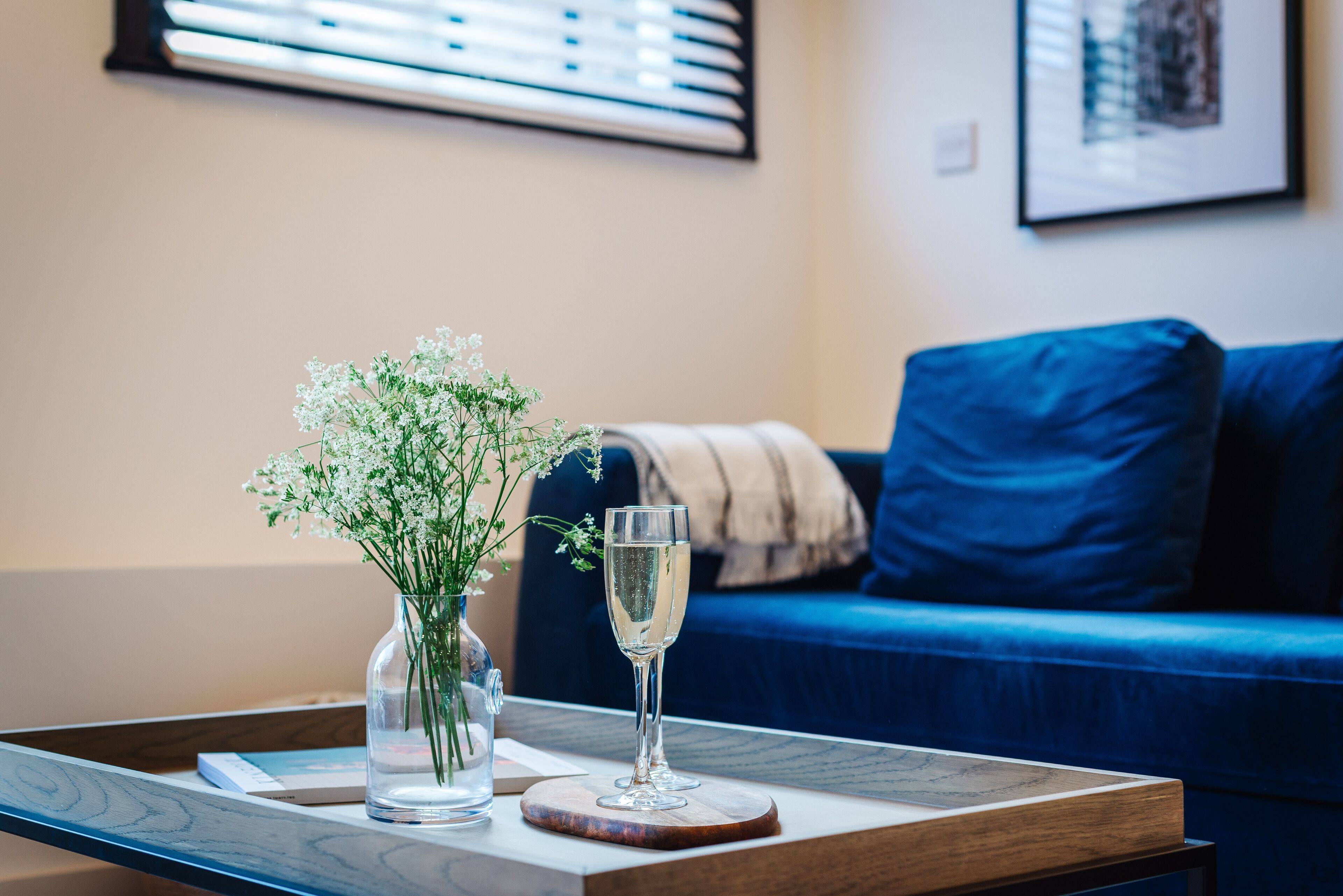 Ein Glas Wasser auf einem Holzteller, eine Vase mit weißen Blumen und ein blaues Sofa im Hintergrund.