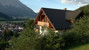 Ein Berghaus mit geneigtem Dach, einer Holzbalkon und Blick auf ein Tal und Berge im Hintergrund.