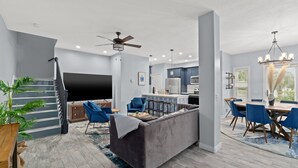 A modern living room with a grey sofa, blue chairs, a dining area with a round table, and a kitchen in the background. - Newly- updated home, a block away from the beach! (Miramar Beach)