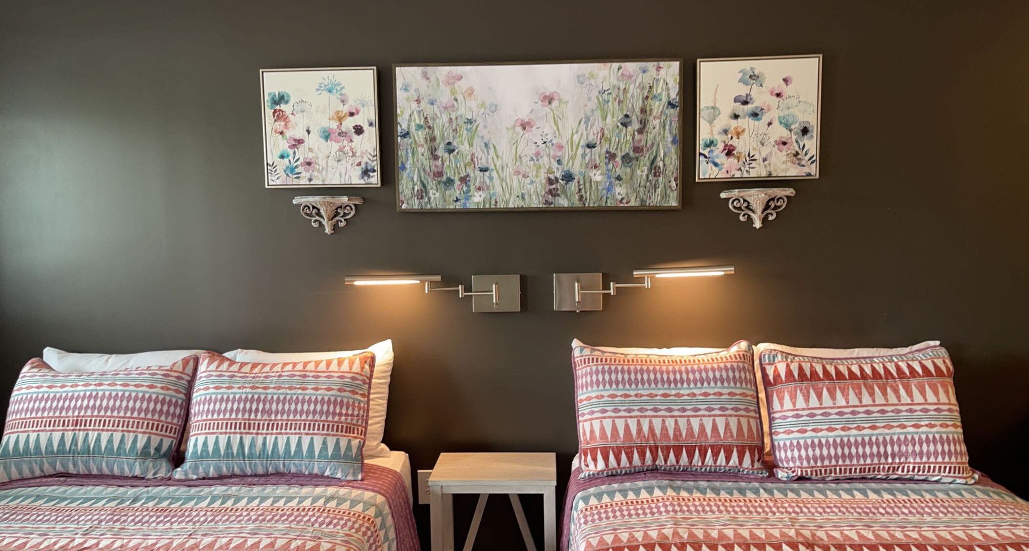 Une chambre avec deux lits, chacun agrémenté de draps à motifs, une petite table de chevet et des appliques murales. Au-dessus des lits, on peut voir trois œuvres d’art florales encadrées.