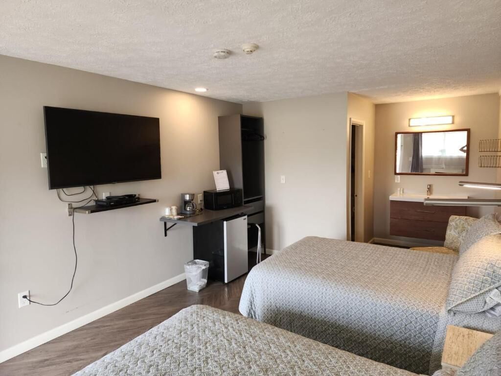 A hotel room with a flat-screen TV mounted on the wall, a kitchenette with a refrigerator and microwave, a bed with patterned bedding, and a mirror above the sink.