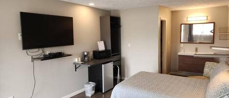 A hotel room with a flat-screen TV mounted on the wall, a kitchenette with a refrigerator and microwave, a bed with patterned bedding, and a mirror above the sink.