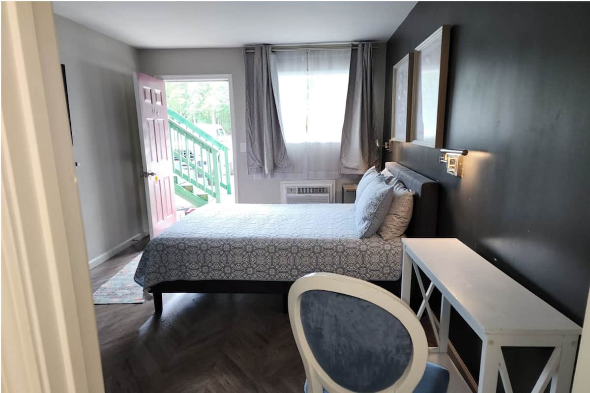 A bedroom with a bed, a desk, a chair, a window with curtains, and a view of a staircase.