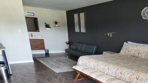 Un dormitorio moderno con una cama grande, suelo de madera oscura, un sofá gris, un baño pequeño y un espejo montado en la pared.