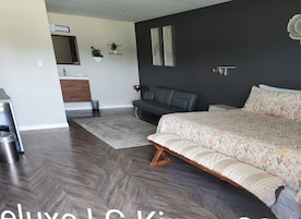 Un dormitorio moderno con una cama grande, suelo de madera oscura, un sofá gris, un baño pequeño y un espejo montado en la pared.