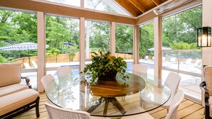 A glass-enclosed patio with a round table and wicker chairs, overlooking a backyard with a pool and lounge chairs. - (2027 OPEN!) Gorgeous Grand Beach Home With Private Pool! (Grand Beach)