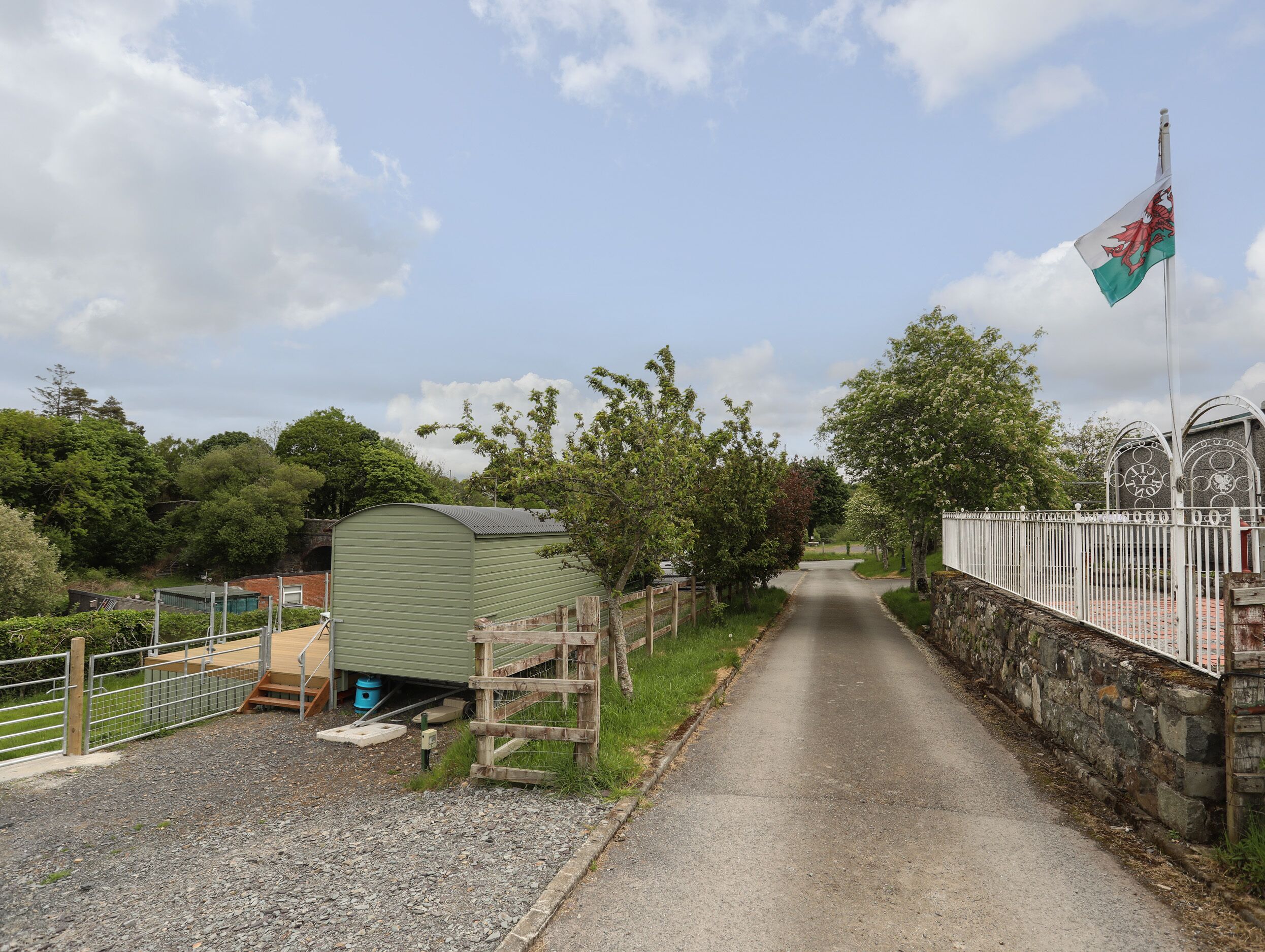 A narrow rural road flanked by a metal fence and a stone wall, leading to a green building with a metal roof.