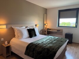 Habitación de hotel con cama, lámparas de noche, escritorio, silla, radiador y ventana con vistas.