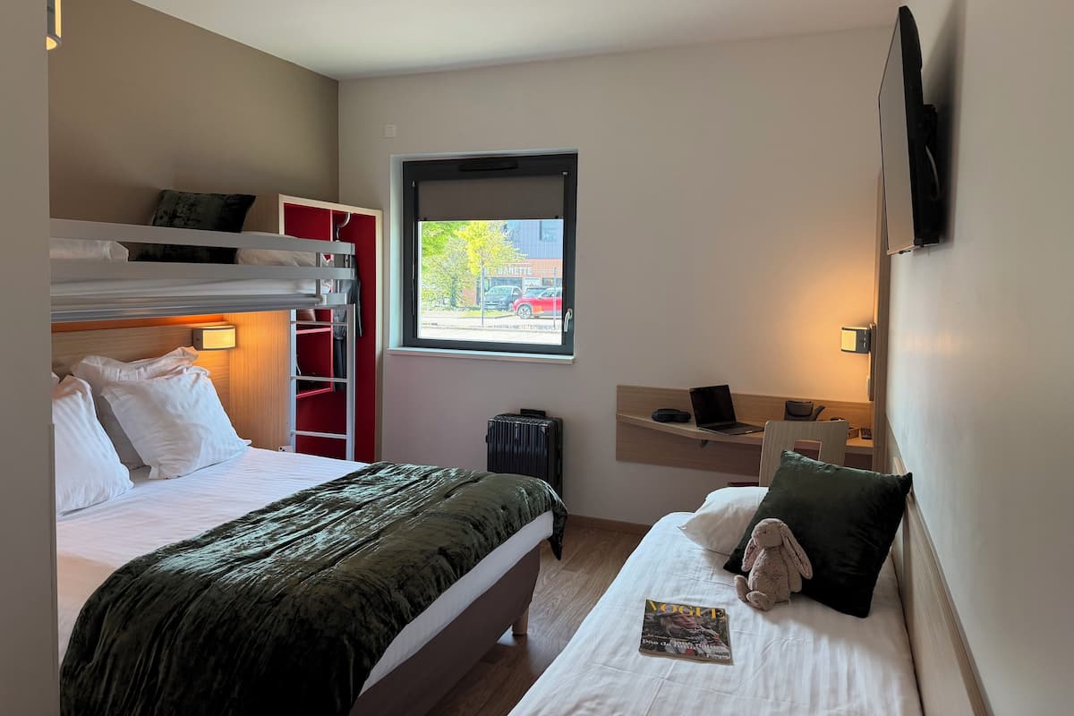 A hotel room with a bunk bed, a desk with a laptop, a window with a view of a building, and a bedside table with a lamp.