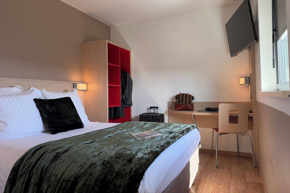 A hotel room with a bed, a desk, a chair, a wardrobe, and a TV mounted on the wall.