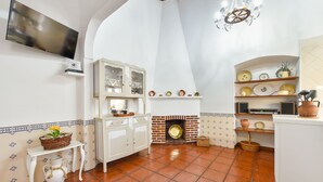 A kitchen with a tiled floor, a white cabinet, a television, a fireplace, and shelves with dishes.
