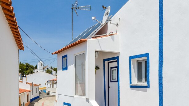 A narrow street with white buildings painted in blue and red accents, a clear blue sky, and a satellite dish on one of the buildings.