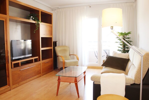 A modern living room with a glass coffee table, wooden shelving, a flat-screen TV, a comfortable sofa, and a lamp.
