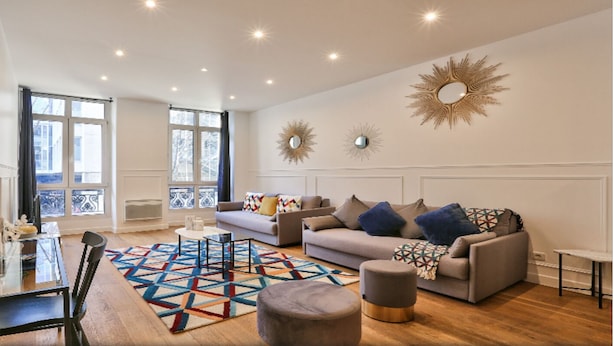 A spacious living room with a sofa, ottoman, and coffee table, a dining area with chairs and a table, and large windows with curtains. - Palais Royal – Musée du Louvre (line 1 and line 7), Pyramides & Bourse (line 3). (Paris)