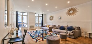 Amplia sala de estar con sofá, reposapiés y mesa de centro, un comedor con sillas y mesa, y ventanas grandes con cortinas.