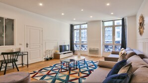 A modern living room with a sofa, a coffee table, and a TV. There are large windows and a balcony. - Palais Royal – Musée du Louvre (line 1 and line 7), Pyramides & Bourse (line 3). (Paris)