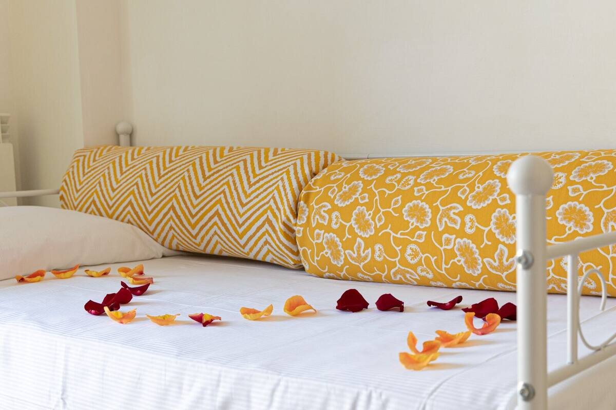 Un lit avec une literie blanche et des coussins à motifs jaunes, une tête de lit en métal blanche et des pétales de rose éparpillés.