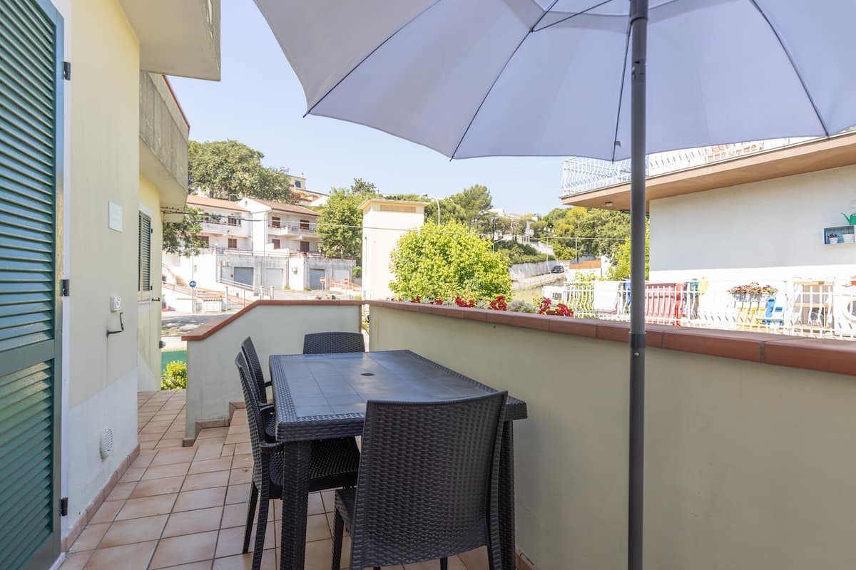 Un balcon avec une table et des chaises, un grand parasol, et une vue sur les bâtiments et la végétation environnants.