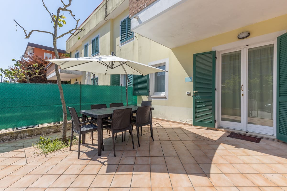 Une terrasse extérieure avec une table et des chaises, une clôture verte et un bâtiment avec un mur jaune et des volets verts.