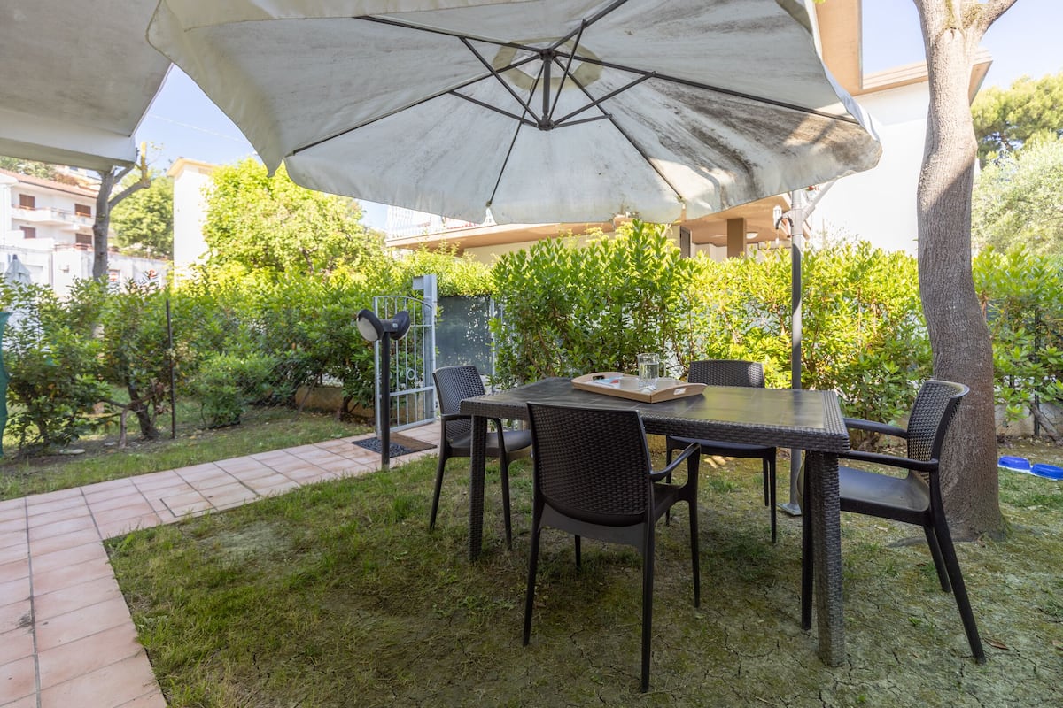Une terrasse extérieure avec une table et des chaises sous un grand parasol, entourée de verdure et bordée d’un chemin en briques.