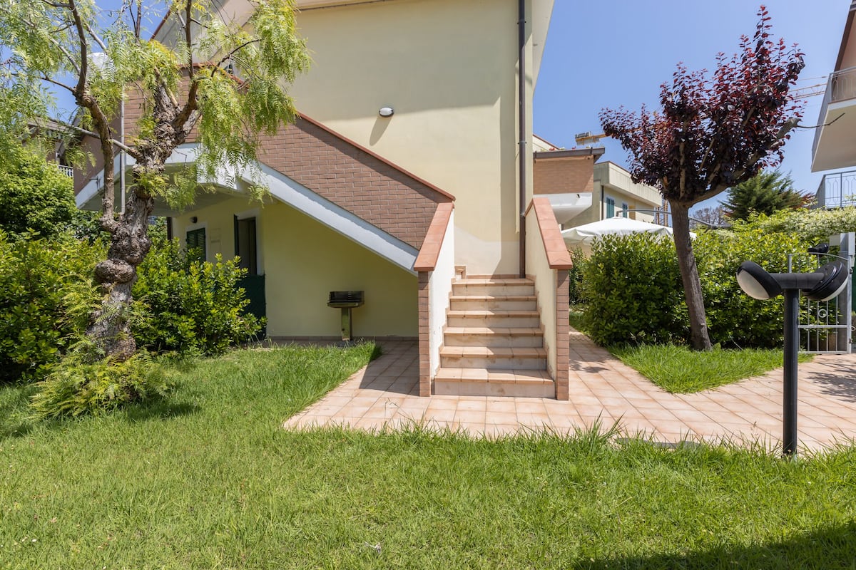 Un immeuble d’habitation avec un escalier menant à une porte, entouré de verdure et sous un ciel dégagé.
