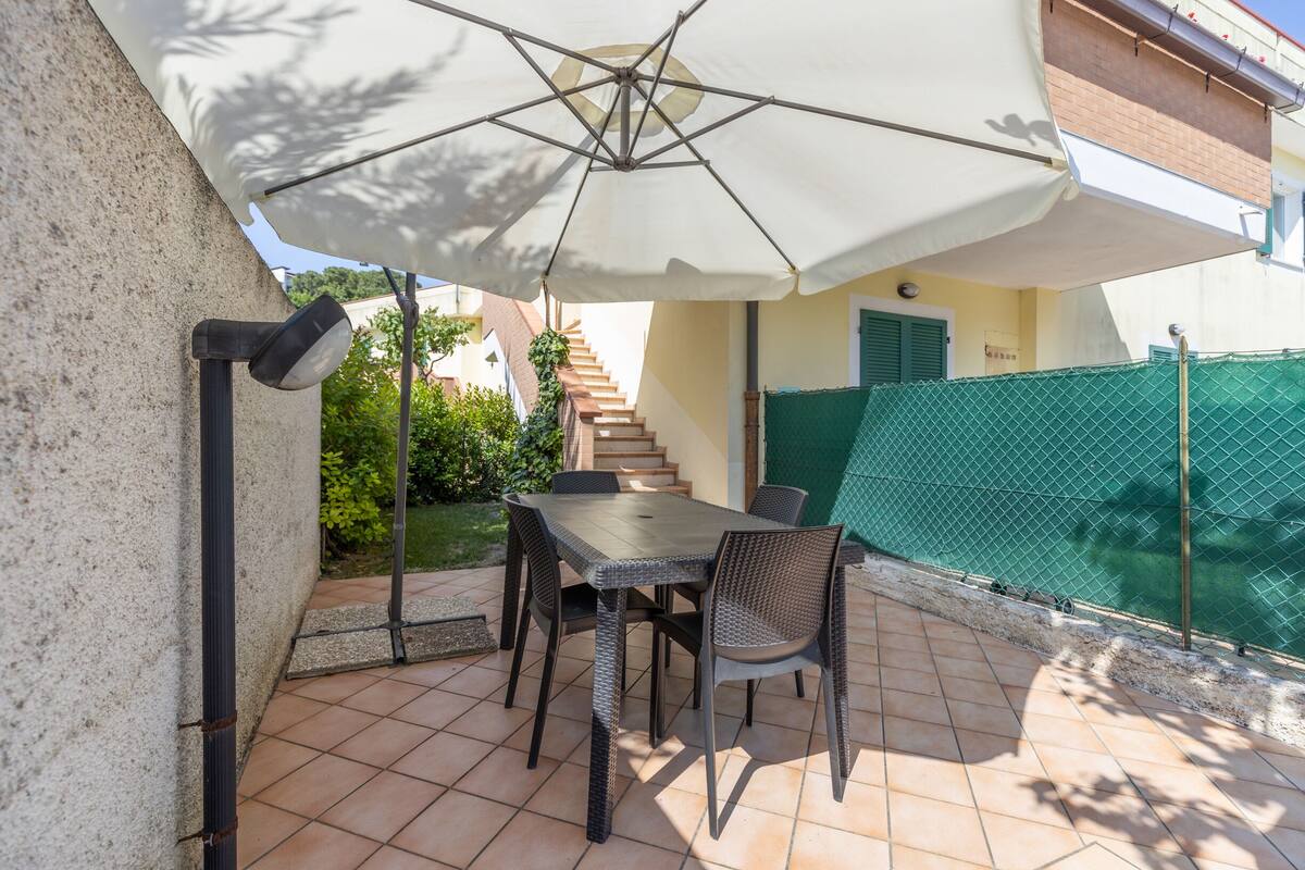 Une terrasse extérieure avec une table et des chaises, un grand parasol et une clôture verte en arrière-plan.
