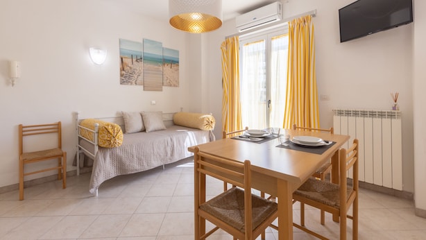 Una habitación con cama, una mesa de comedor con sillas, una lámpara colgante, un televisor de pantalla plana montado en la pared y una ventana con cortinas amarillas.