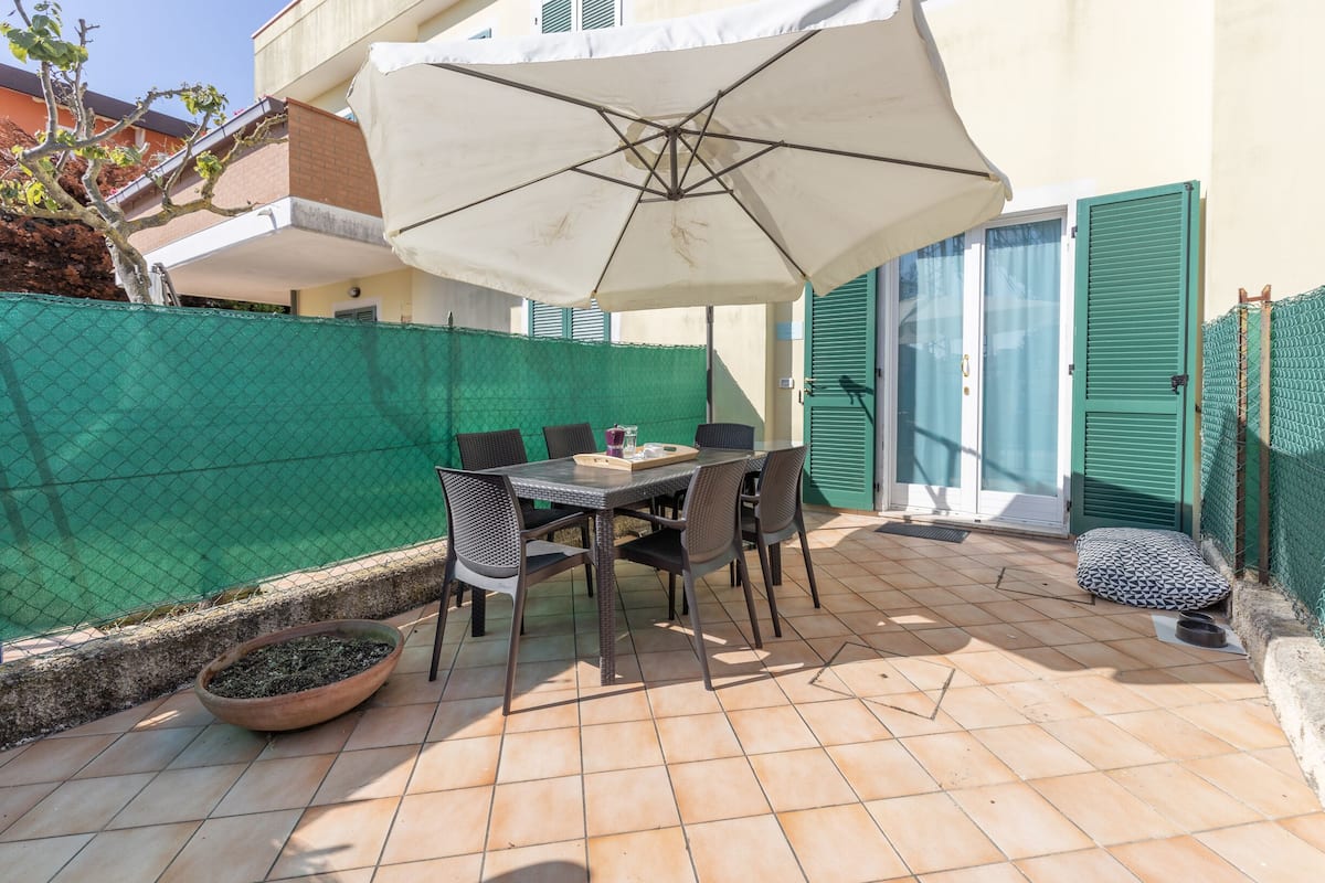 Une terrasse extérieure avec une table et des chaises, un grand parasol et une clôture verte.