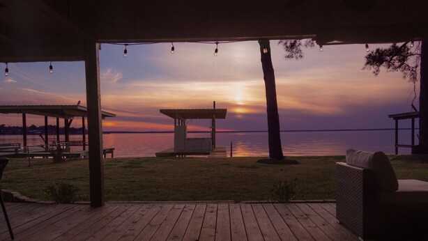 Eine Holzterrasse mit Blick auf den Sonnenuntergang über dem Wasser, ein überdachter Sitzbereich und ein kleines Pavillon.