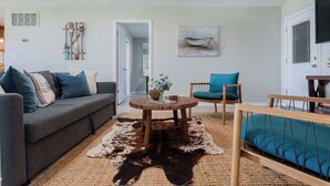 Ein Wohnzimmer mit grauem Sofa, einem runden Holztisch und einem türkisfarbenen Sessel. Ein Kuhfellteppich bedeckt den Boden.