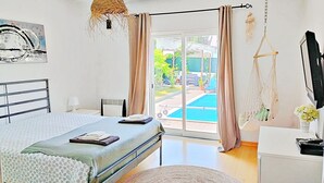A bedroom with a bed, a desk, a chair, a hanging chair, and a view of a pool and garden. - A Casa da Lagoa (Lagoa de Albufeira) - 800 da Praia (Sesimbra)