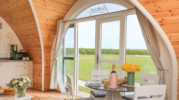 Une pièce chaleureuse avec un plafond en bois, une grande fenêtre, une table à manger avec des chaises, un petit coin cuisine et une vue sur un champ verdoyant.