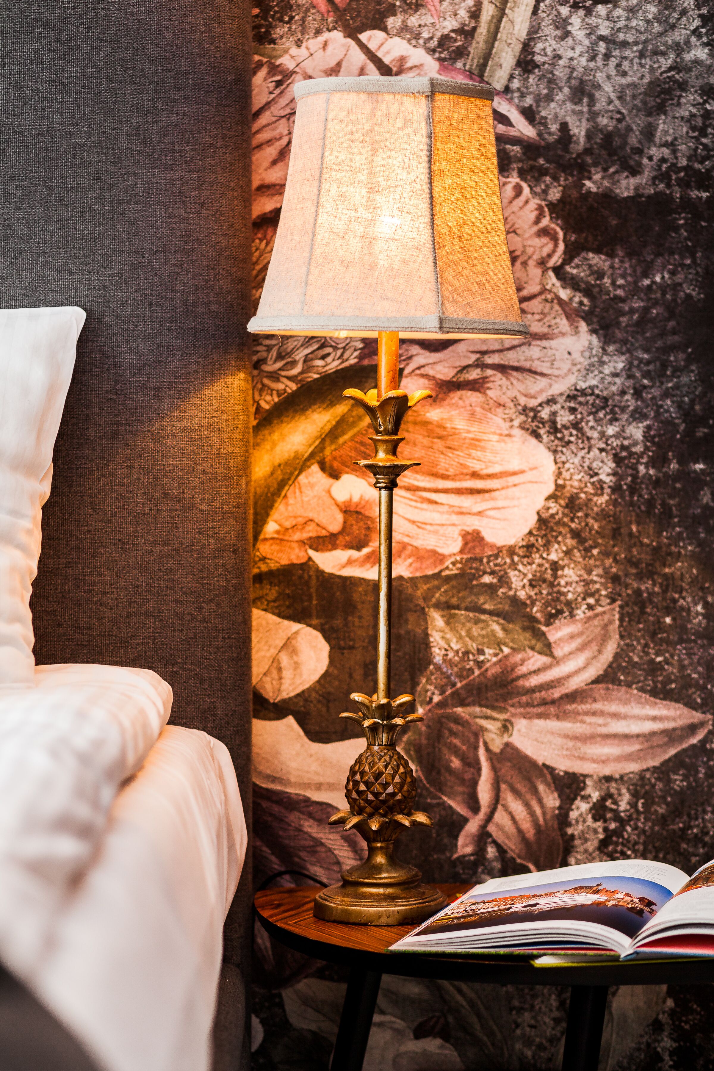 Una cama con edredón blanco, una lámpara con pantalla color crema y un libro sobre una mesita de noche.