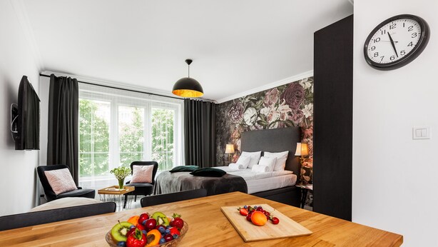 Quarto de hotel com uma mesa de jantar de madeira, uma cesta de frutas, uma tábua de corte, uma cama com cabeceira, um relógio de parede e uma janela com cortinas.