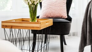 Quarto moderno com uma cadeira preta, uma mesinha de madeira com um vaso de flores brancas e uma almofada rosa.