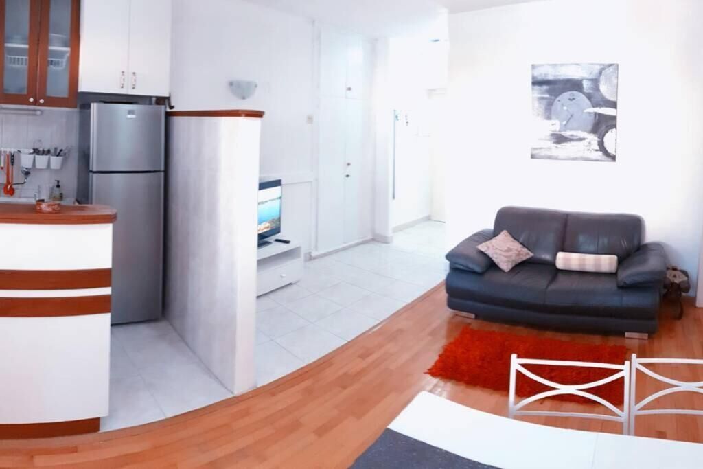 Een moderne keuken met witte kasten, een roestvrijstalen koelkast en een houten aanrecht. In de woonkamer staat een grijze bank met een rood tapijt.