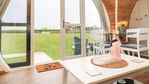 Une pièce moderne dotée d’une grande porte vitrée donnant sur un espace extérieur verdoyant. On y trouve une table blanche, sur laquelle sont posés une bouteille et un vase de fleurs.