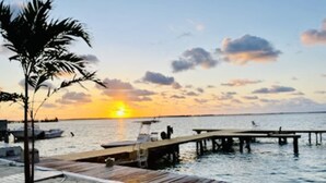 Une jetée en bois s’étendant sur des eaux calmes au coucher du soleil, avec un palmier et un bateau amarré à proximité.