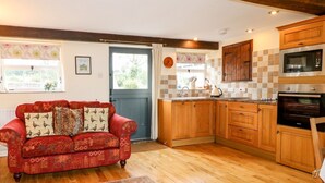Un salon chaleureux avec un canapé rouge et des coussins à motifs, un plancher en bois et une cuisine dotée de meubles en bois et d’appareils électroménagers modernes.