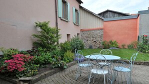 Une cour avec une table et des chaises, entourée d’un jardin où poussent différentes plantes et fleurs.