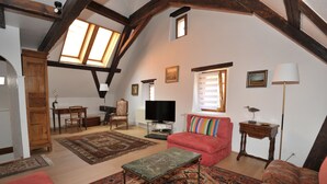 Un salon spacieux avec un plafond haut, des poutres en bois, un téléviseur, un canapé rouge, un tapis à motifs et une petite table.