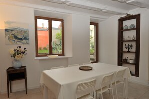A dining area with a table set for four, a window with a view of a building and greenery, and a shelf with decorative items.