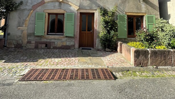 Une maison traditionnelle en pierre, dotée d’un toit en pente, d’une entrée centrale avec une porte en bois et de deux fenêtres avec volets.