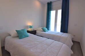Two single beds with white duvets and teal pillows, a nightstand with a lamp, and a window with blue curtains.