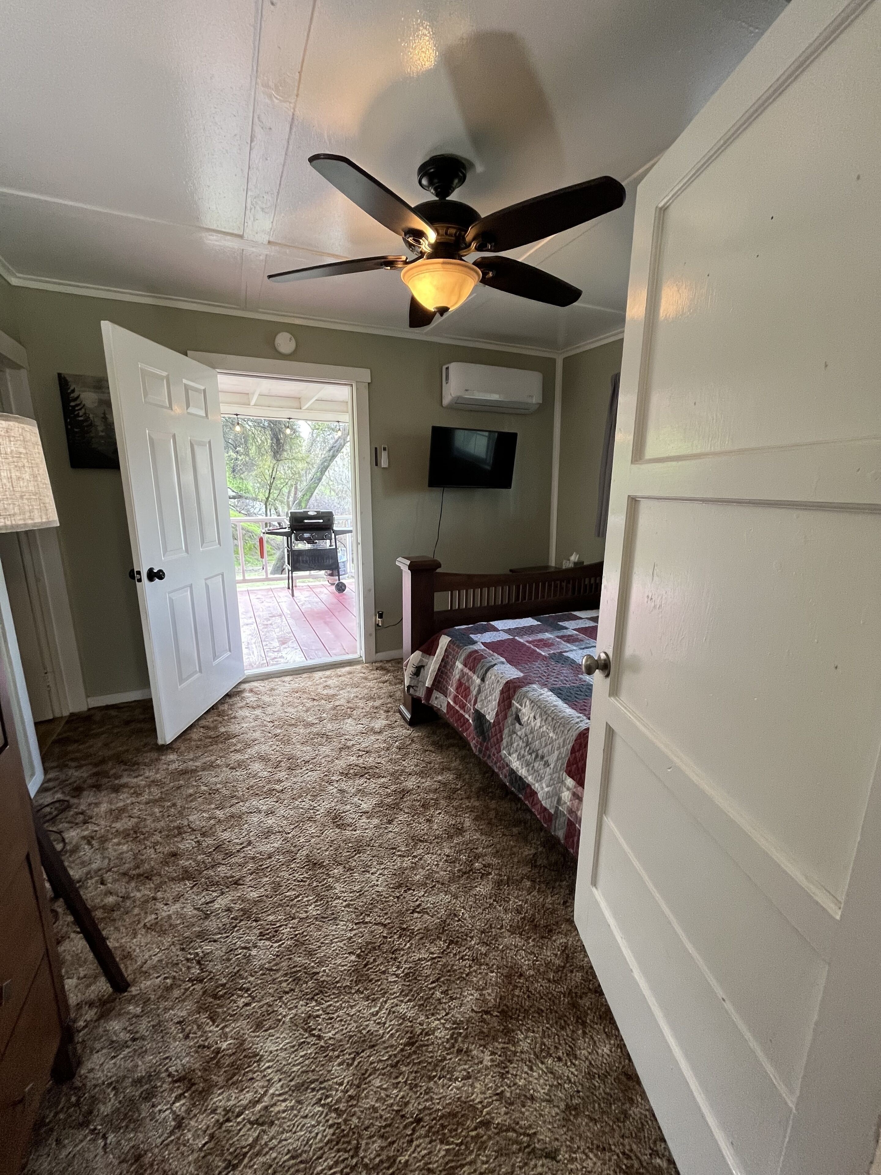 Un dormitorio con cama, ventilador de techo, televisor y puerta que da a un área exterior.