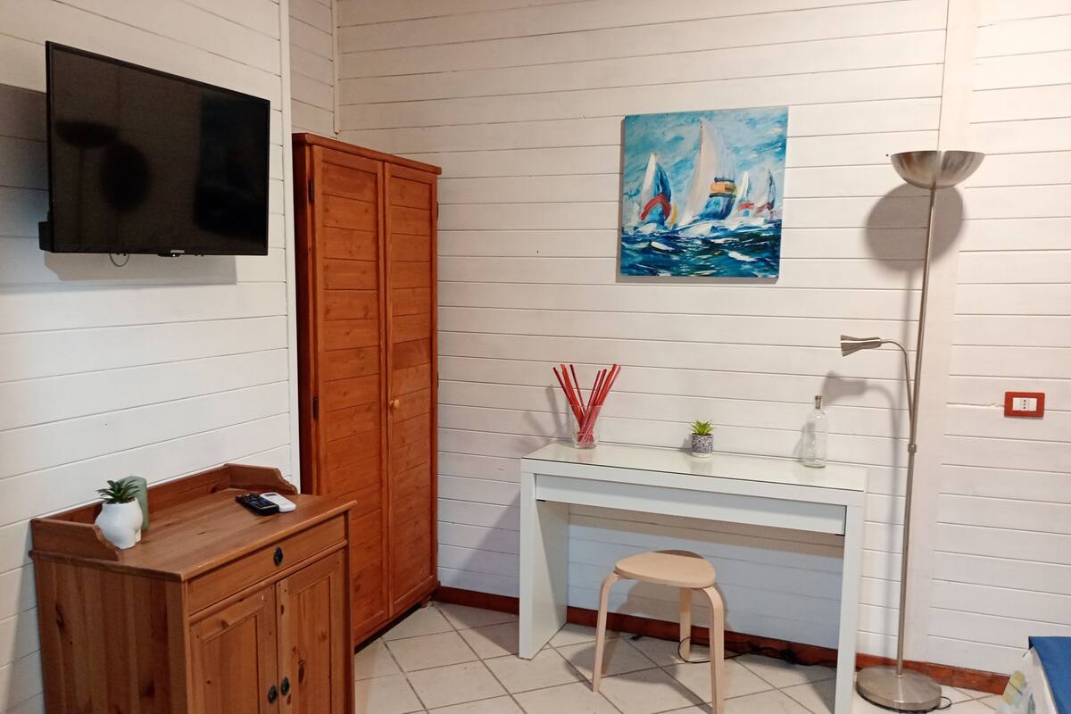 A room with a wooden cabinet, a desk with a stool, a painting of sailboats, and a floor lamp.