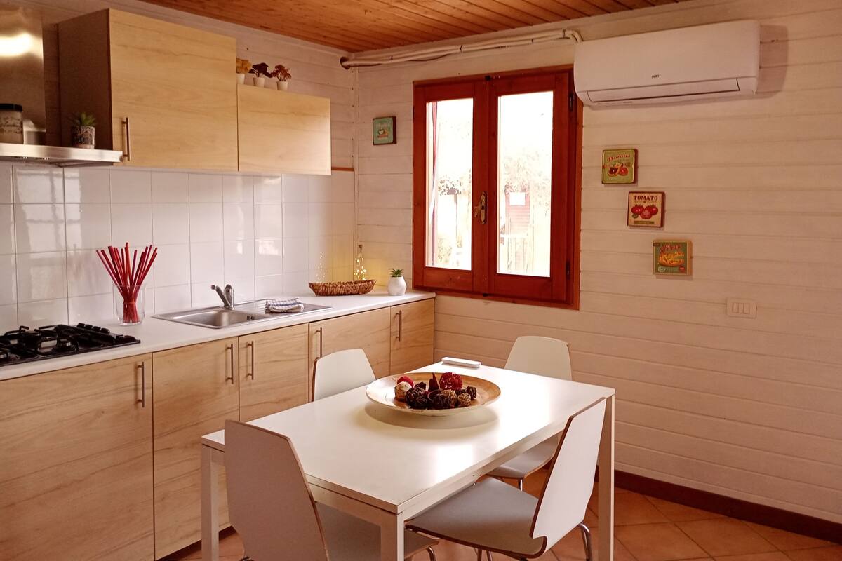 A kitchen with wooden cabinets, a white countertop, a dining table with chairs, and a window with red frames.