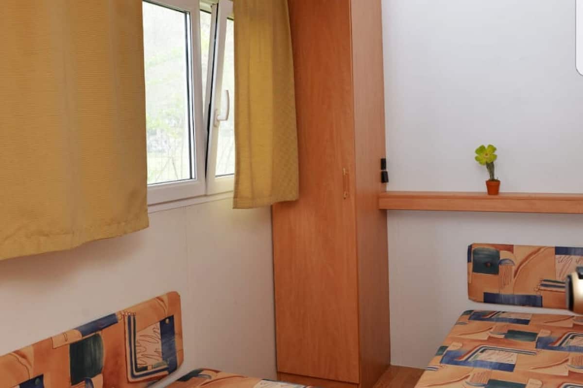 A room with two beds, a wooden headboard, a small shelf with a plant, and a window with curtains.