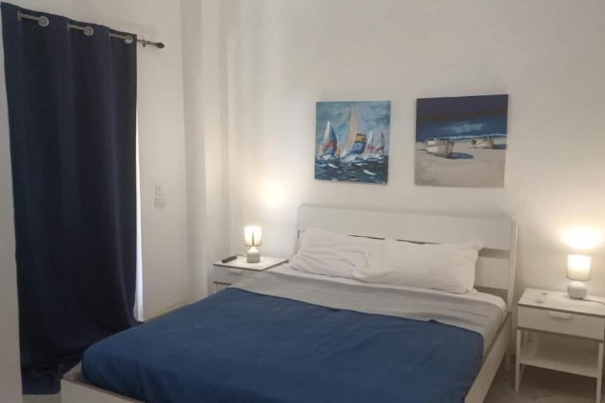 A bedroom with a bed, two bedside tables with lamps, a dark blue curtain, and two paintings on the wall.